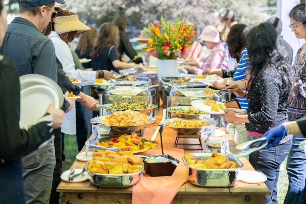A long buffet table laden with various dishes is being served by a diverse group of people outdoors.
