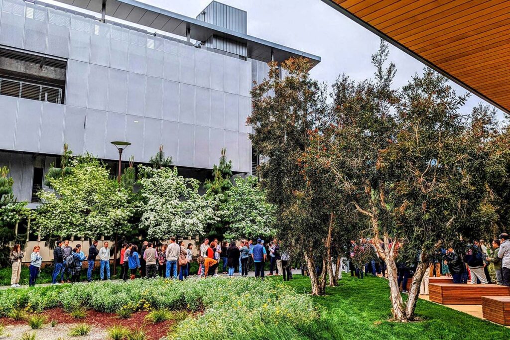 A long line of people waits outside a modern building on the Stanford University Redwood City campus, in a landscaped area with trees and overcast skies.