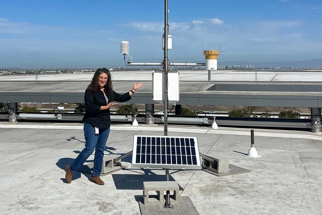 A woman stands on a rooftop next to a weather station powered by a solar panel, gesturing towards the equipment under a clear sky.