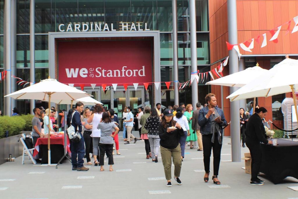 A crowd of people gathers outside Cardinal Hall at Stanford University, with white umbrellas, tables, and "We ❤️ Stanford" signage visible.
