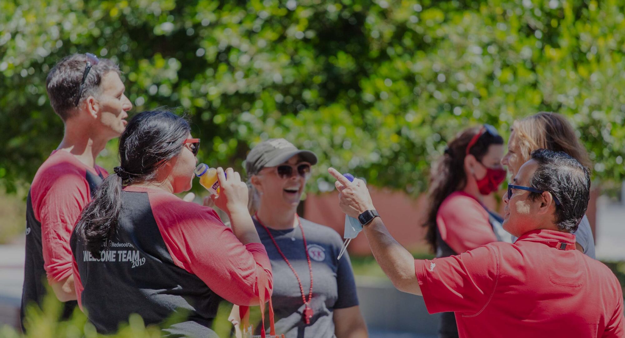 Six adults wearing matching red and black shirts stand outdoors, interacting and drinking from water bottles.