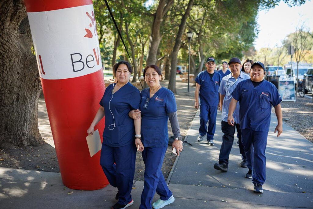 A group of healthcare workers in blue scrubs walk and smile together along a sun-dappled outdoor path next to a large red and white inflatable pillar.