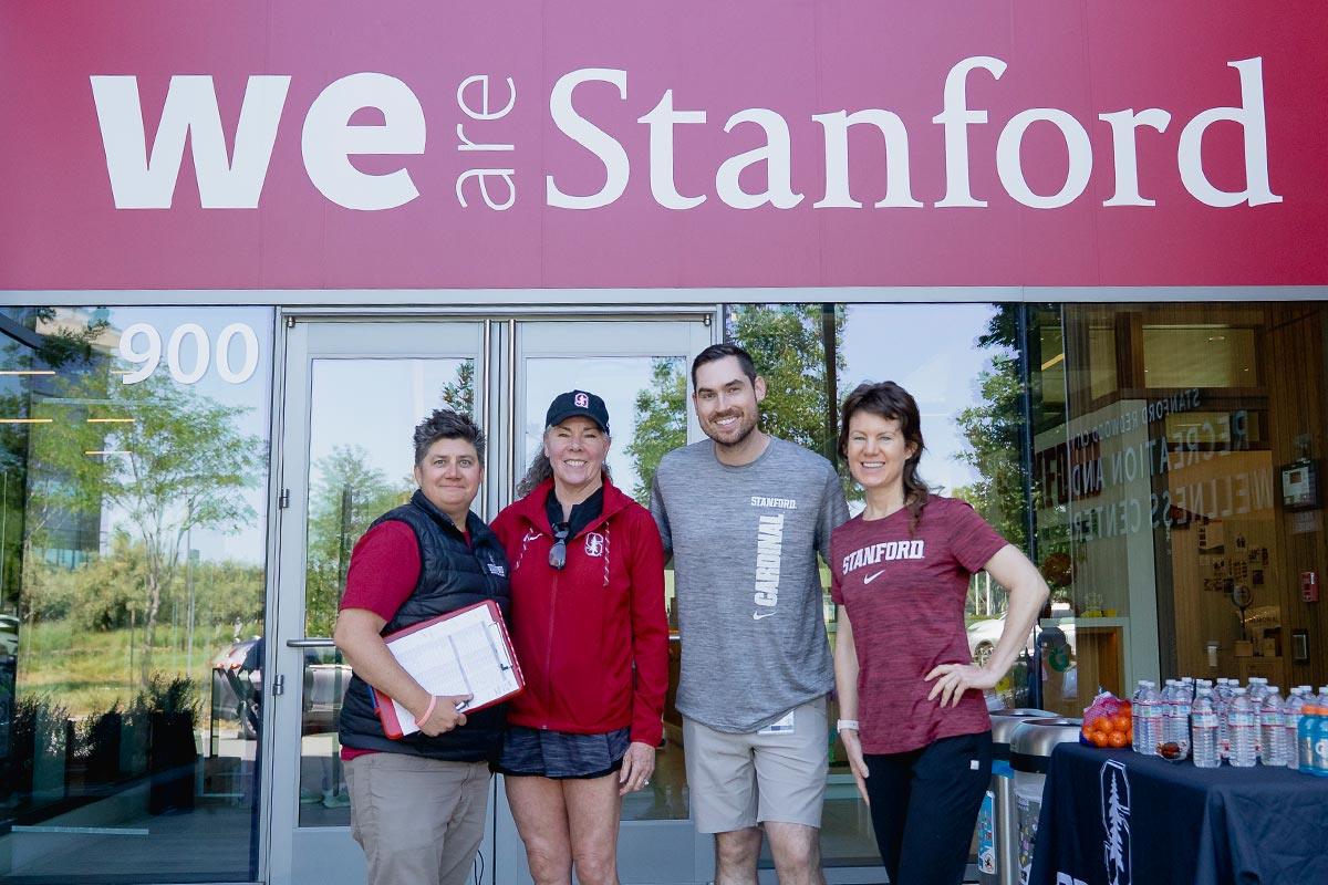 Four Stanford staff members in university-branded athletic gear smile next to a table with water bottles and oranges.
