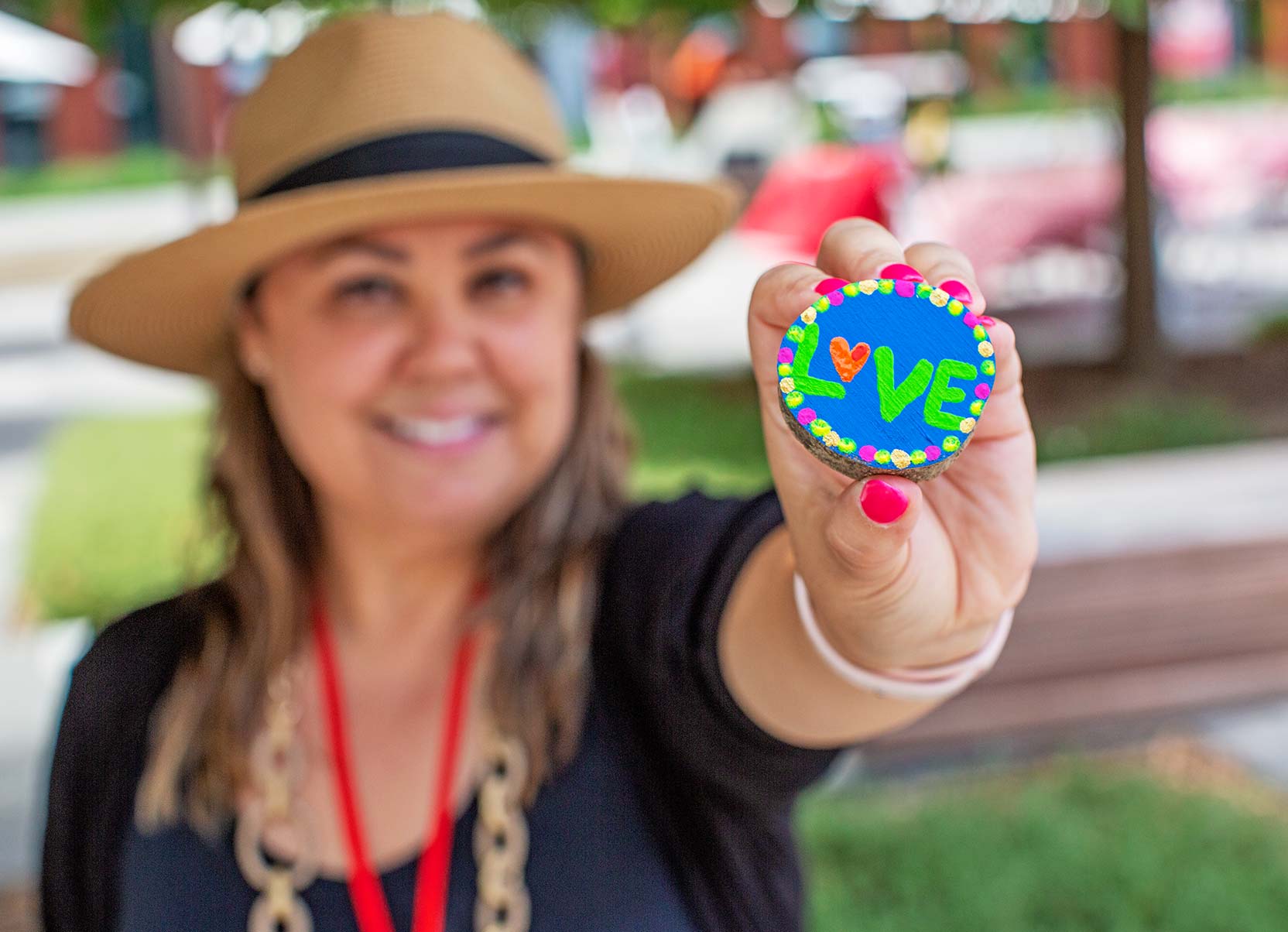 A smiling woman in a tan hat holds up a small, hand-painted wooden disc featuring the word "LOVE" in vibrant green lettering.