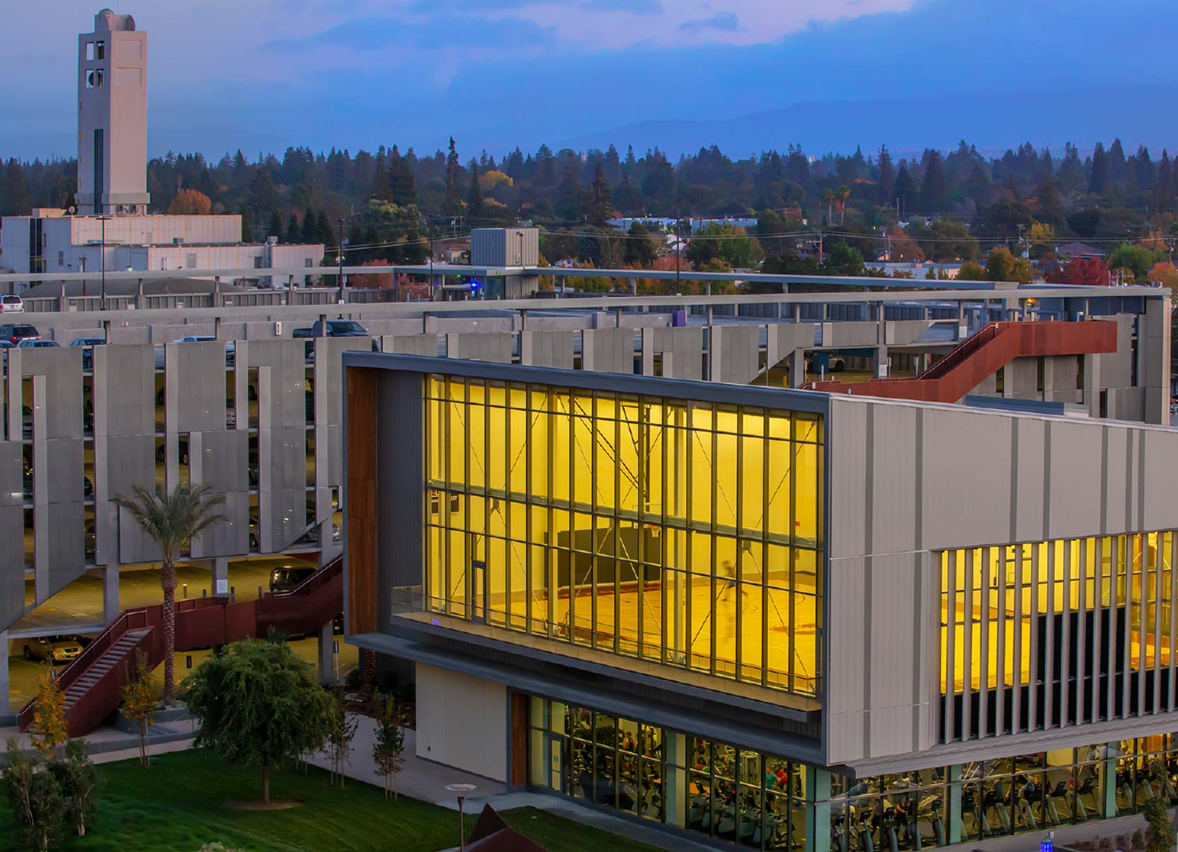 An illuminated, modern multi-story recreation center at Stanford University’s Redwood City campus with large glass windows revealing an indoor basketball court and gym at dusk.