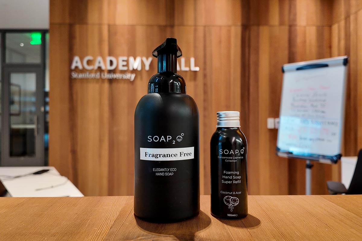 A large, black matte dispenser labeled "SOAP₂ Fragrance Free" sits next to a smaller "Super Refill" bottle on a wooden counter at Stanford University's Academy Hall.