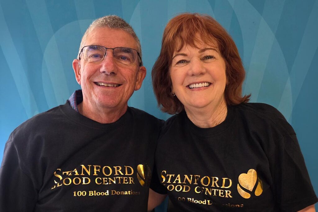 A man and a woman in matching Stanford Blood Center T-shirts smile while holding up a certificate celebrating 100 donations.
