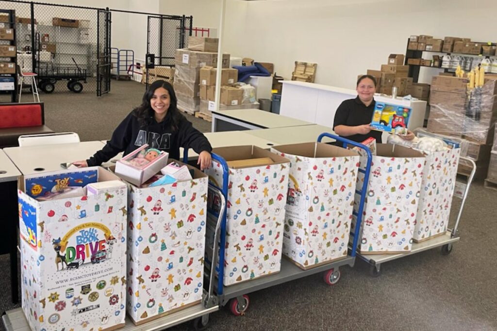 A staff member stands behind a table filled with wrapped gifts and colorful gift bags for the PAL annual Toy & Book Drive.