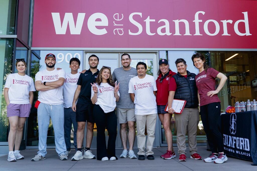 A group of Stanford staff members stands outdoors in front of a building, posing together under a "We Are Stanford" banner.