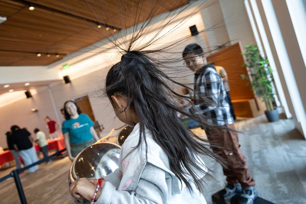 A young girl participates in STEMfest, demonstrating static electricity by touching an electrified metal ball which is making her hair stand up. (Photo by Jim Gensheimer)