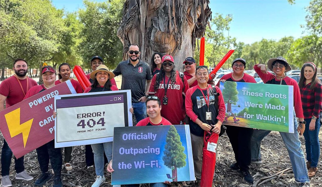 A group of smiling Stanford staff members stand outdoors holding signs and a banner as part of a community outreach event.