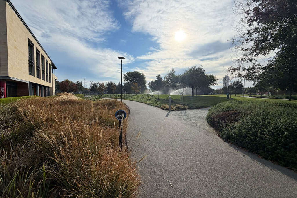 A wide-angle view of the Stanford Redwood City campus features a paved walking path leading toward modern office buildings under a clear sky.