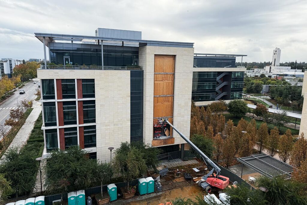 A modern multi-story office building on the Stanford Redwood City campus features large glass windows and an adjacent outdoor patio with native landscaping.